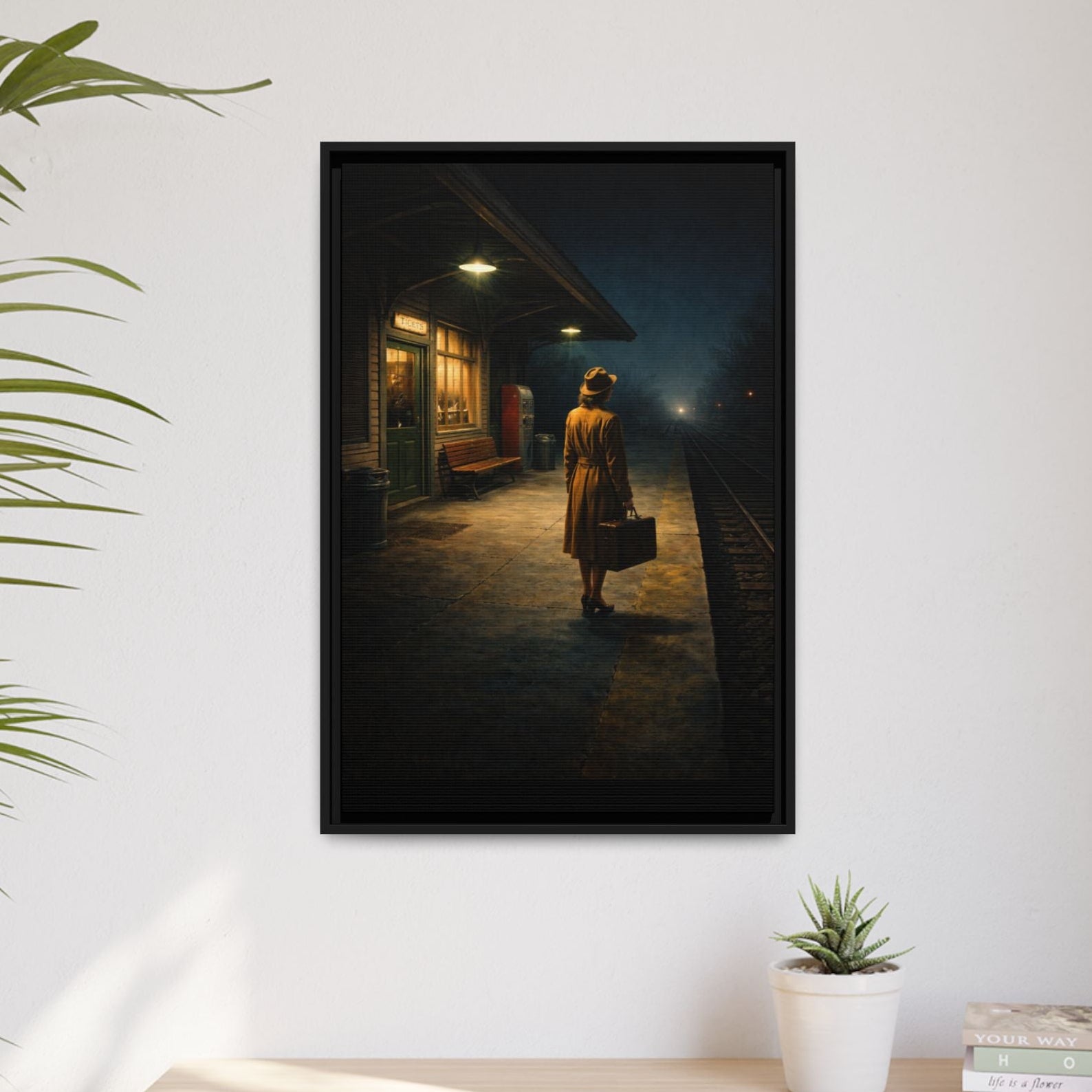 Framed artwork of a woman with a suitcase on a platform, above a desk with a plant and books.